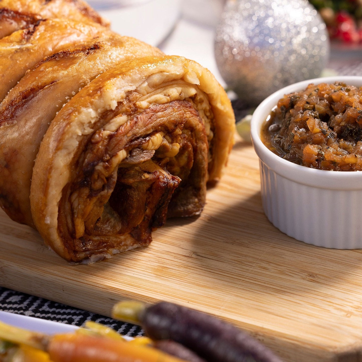 Roasted pork belly roll with crispy crackling sits on a board with apple & sage sauce in a white dish alongside.